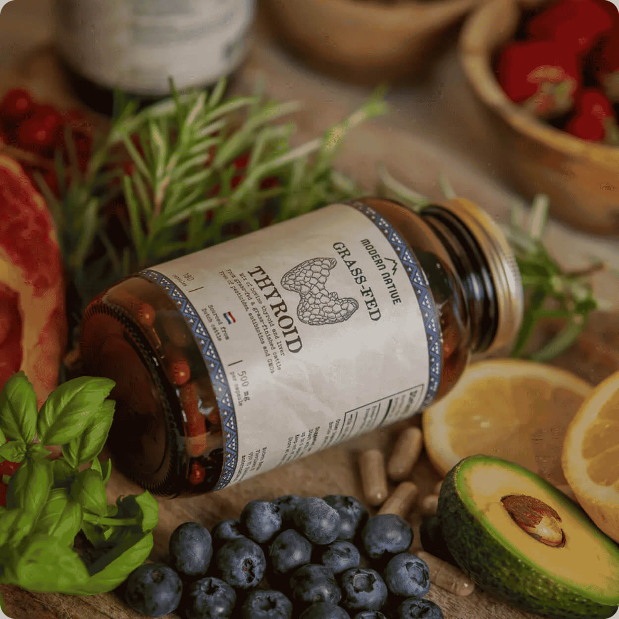 Na dřevěném podkladu leží skleněná dóza s doplňkem stravy Grass-Fed Thyroid, obklopená čerstvými borůvkami, rozkrojeným avokádem, citronem, lístky bazalky, snítkami rozmarýnu a volně sypanými kapslemi.
